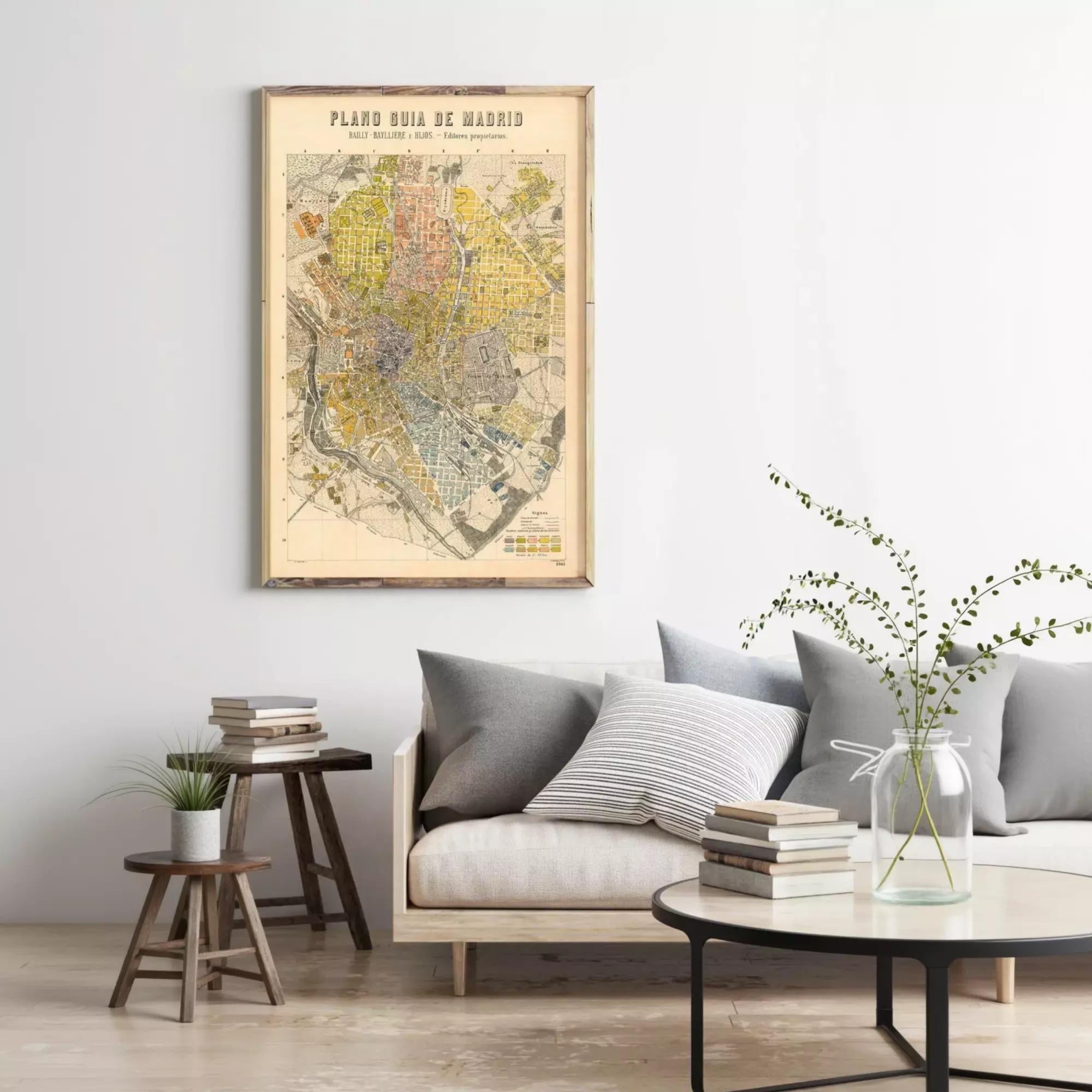 a living room with a framed vintage map of Madrid hanging on the wall above a couch with pillows. The room has a wooden coffee table with a vase of greenery, and there are several books and a plant on the table.