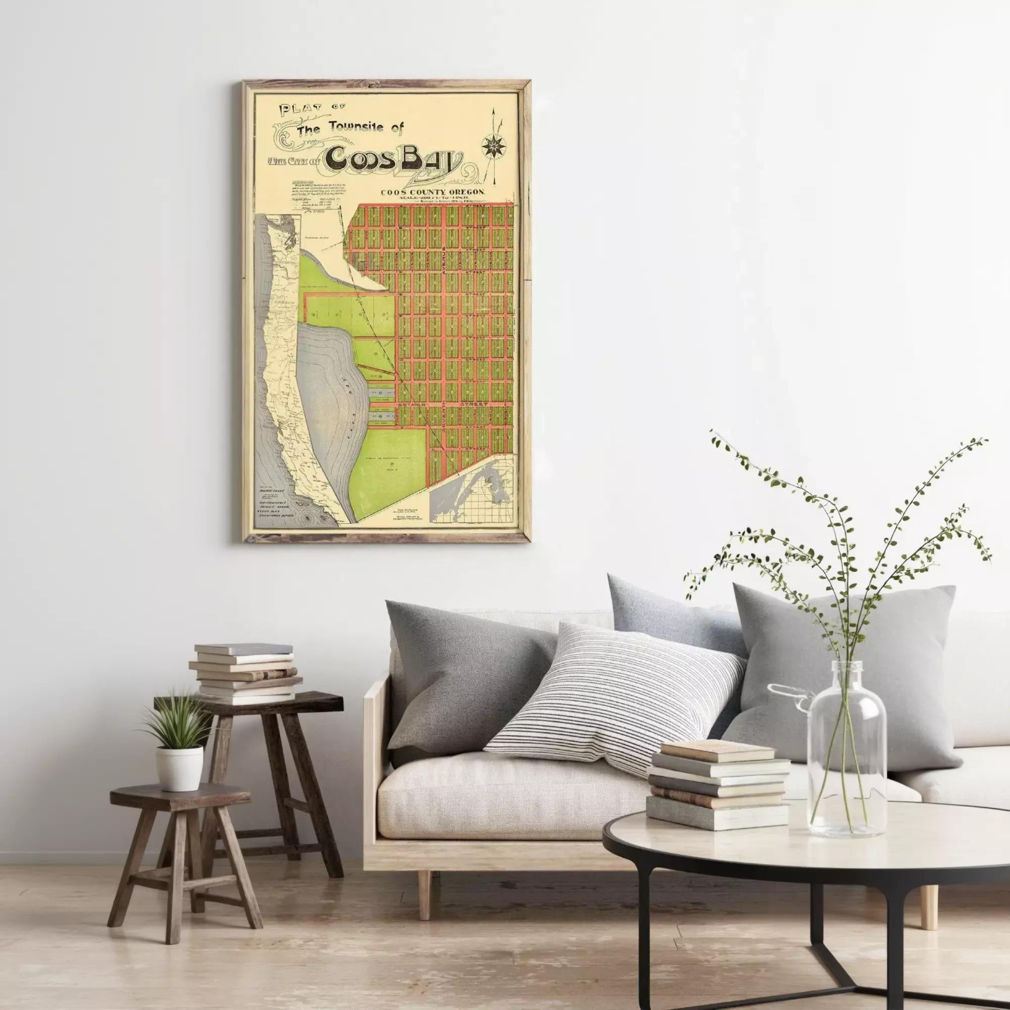 a living room with a framed map of the town of Cos Bay hanging on the wall, a couch with pillows, a coffee table with books and a vase of greenery, and a small table with a plant.