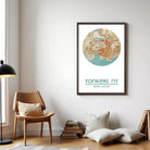 a framed map of New York City hanging on a wall, with a wooden chair and a stack of books in the foreground.