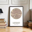 A framed map of London with the text "LONDON 51°32'N 0°57'W" printed on it, placed on a wooden floor next to a stack of books.