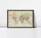 The image is a framed vintage world map with a black border and gold trim, displayed on a white wall.