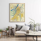 a living room with a large framed map of a city hanging on the wall above a couch with pillows. The room also includes a coffee table with books and a vase with greenery.