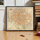 A framed map of the city of Bologna, Italy, displayed on a wooden floor next to a stack of books.
