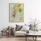 a living room with a large framed map hanging on the wall above a couch with pillows. There are also two small tables with books and plants on them, and a round coffee table in front of the couch.