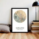 A framed map of Oakland, California, with the text " Oakland 37°48' 12.16 W" printed on it, is displayed on a wall next to a stack of books and a lamp.