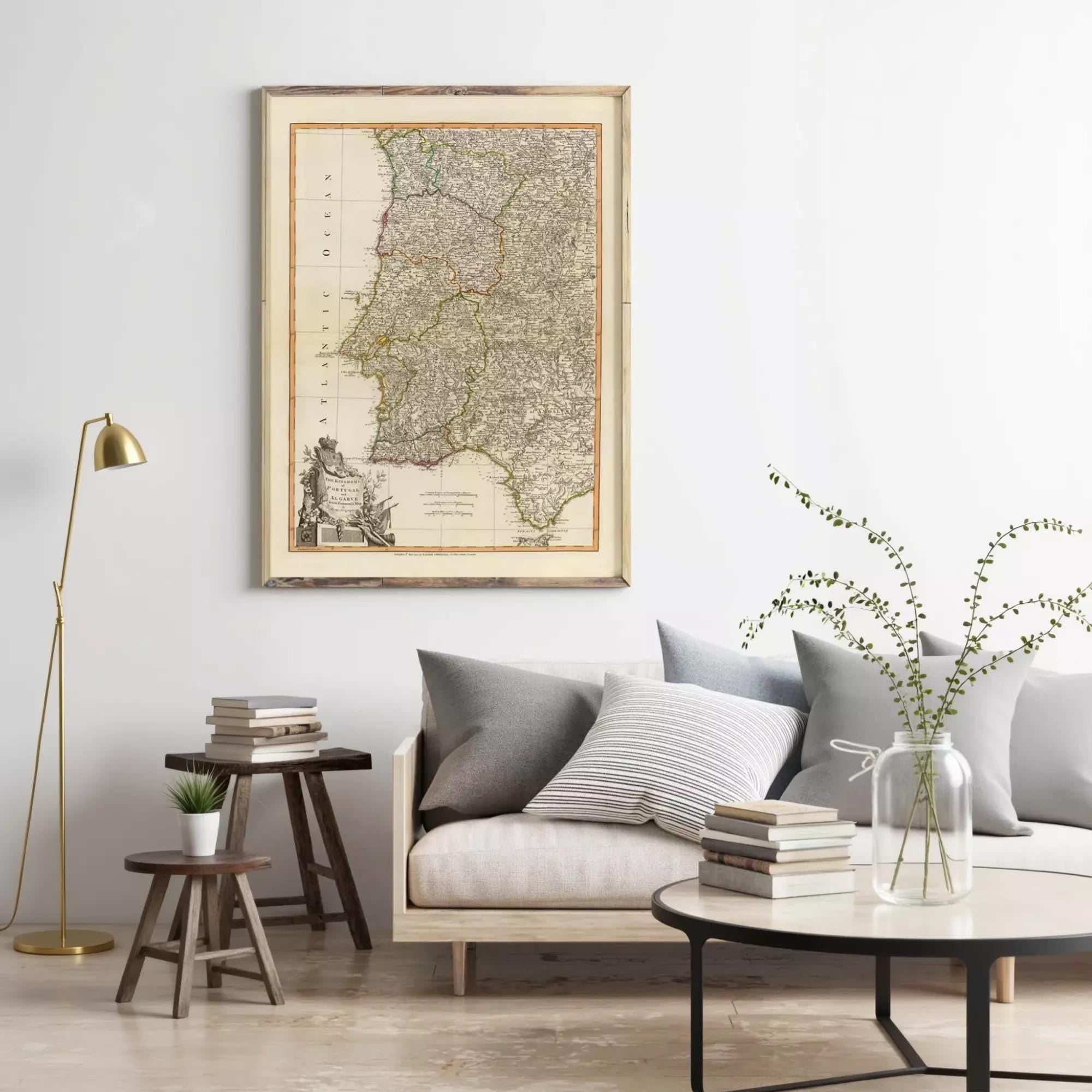 a living room with a large framed map of Portugal hanging on the wall above a gray sofa with striped pillows. The room also includes a wooden side table, a round coffee table with books and a vase of greenery, and a floor lamp.