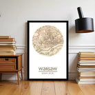 A framed poster of a map of Warsaw, Poland, with the text " Warsaw" and "52.21" below it, is displayed on a wall next to a stack of books and a black lamp.
