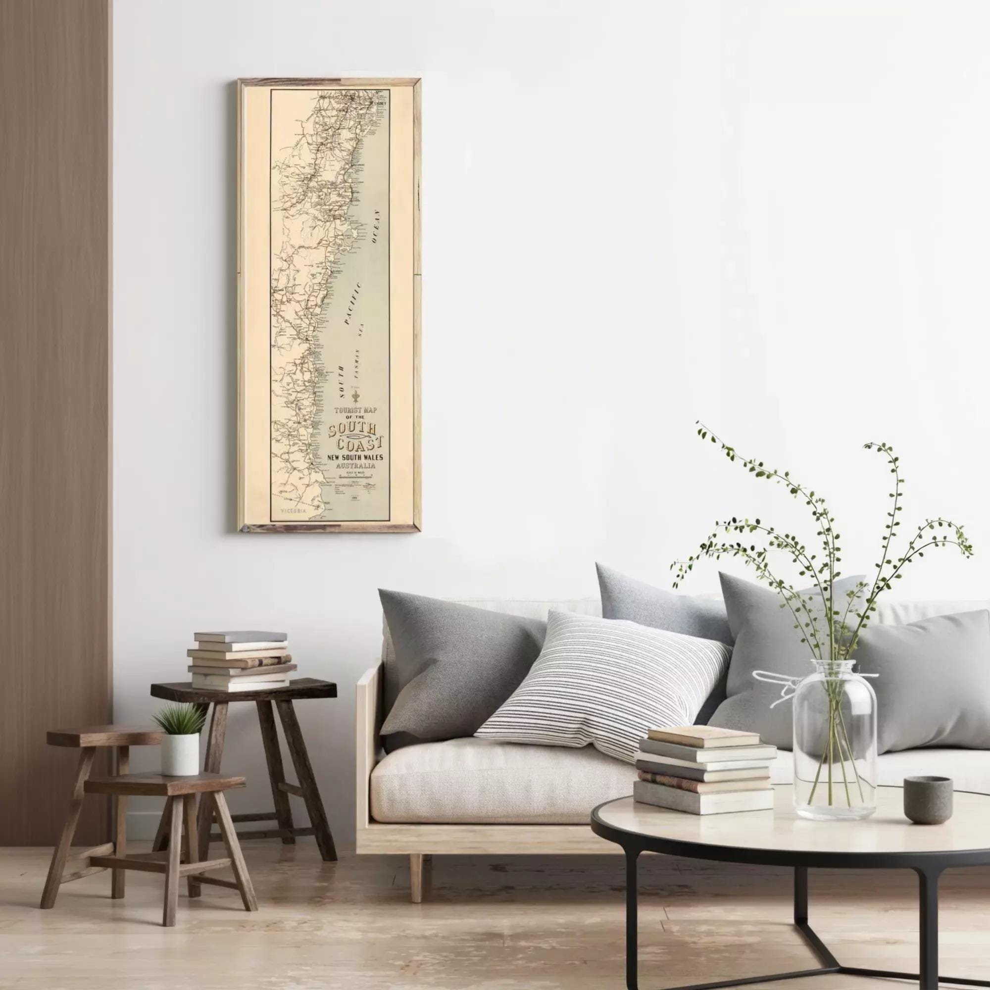 a cozy living room with a large map of the South Coast of Australia hanging on the wall, a comfortable couch with pillows, a coffee table with books and a vase of greenery, and a wooden stool in front of the couch.