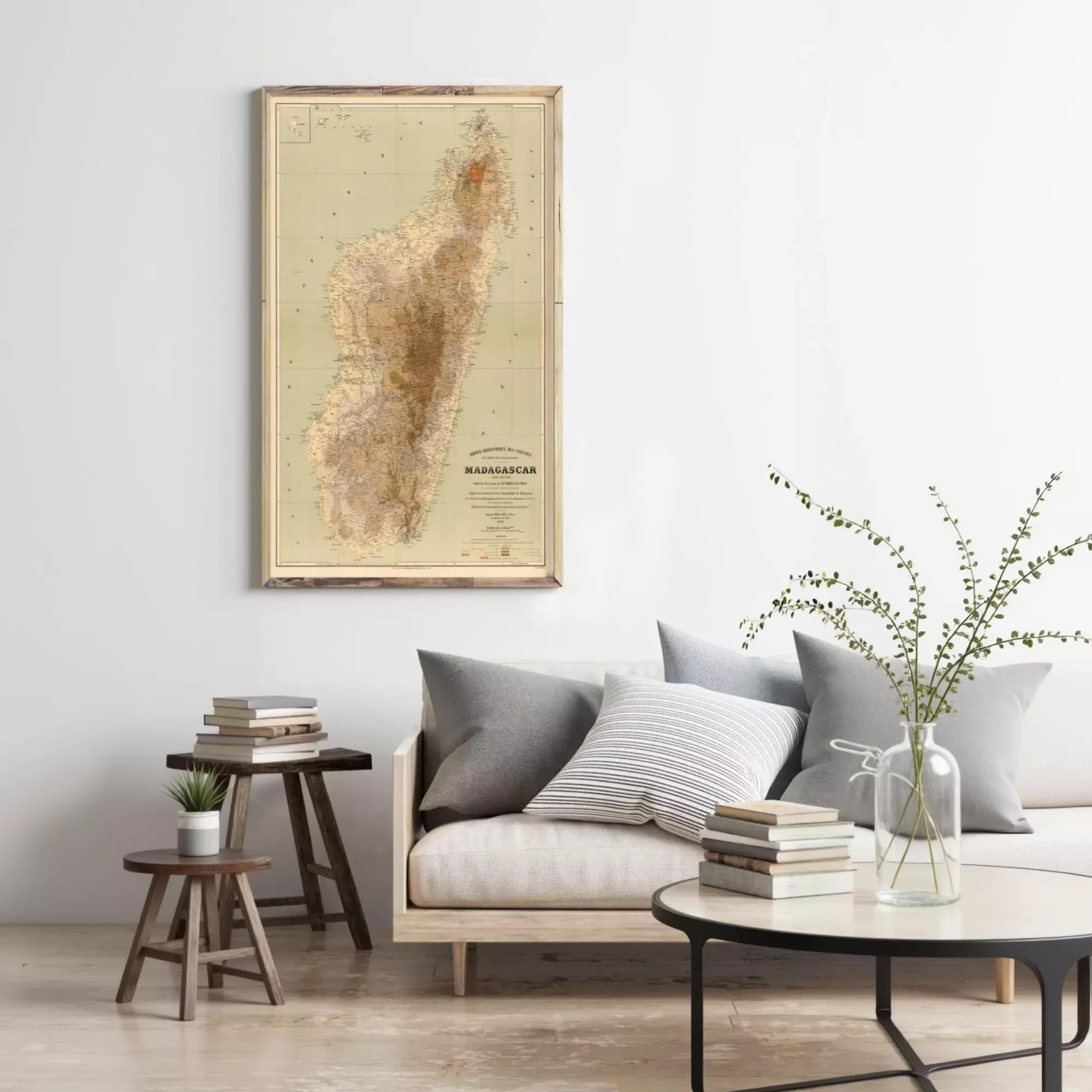 a living room with a large map of Madagascar hanging on the wall, a wooden side table with a plant, a gray sofa with pillows, and a round coffee table with books and a vase of greenery.