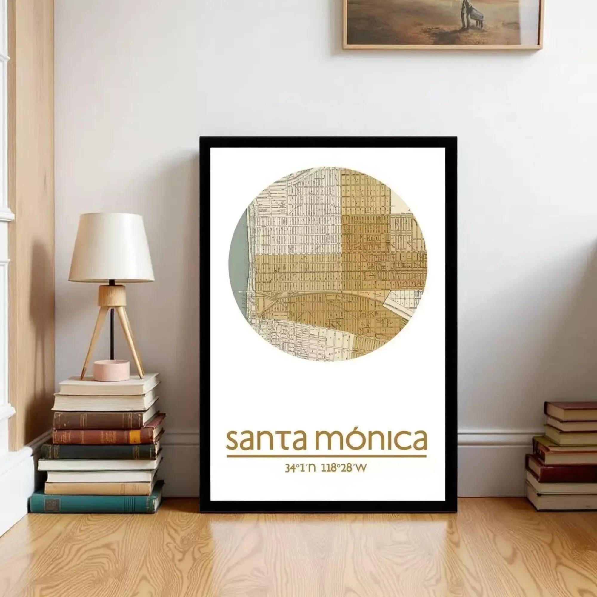 A framed poster of a map of Santa Monica, California, with the text "Santa Monica" and "34° 11' 28" W" printed on it. The poster is displayed on a wall next to a stack of books and a lamp.