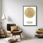 a living room with a framed map of Oxford, England, hanging on the wall. The room has a gray armchair, a wooden side table, and a stack of books.