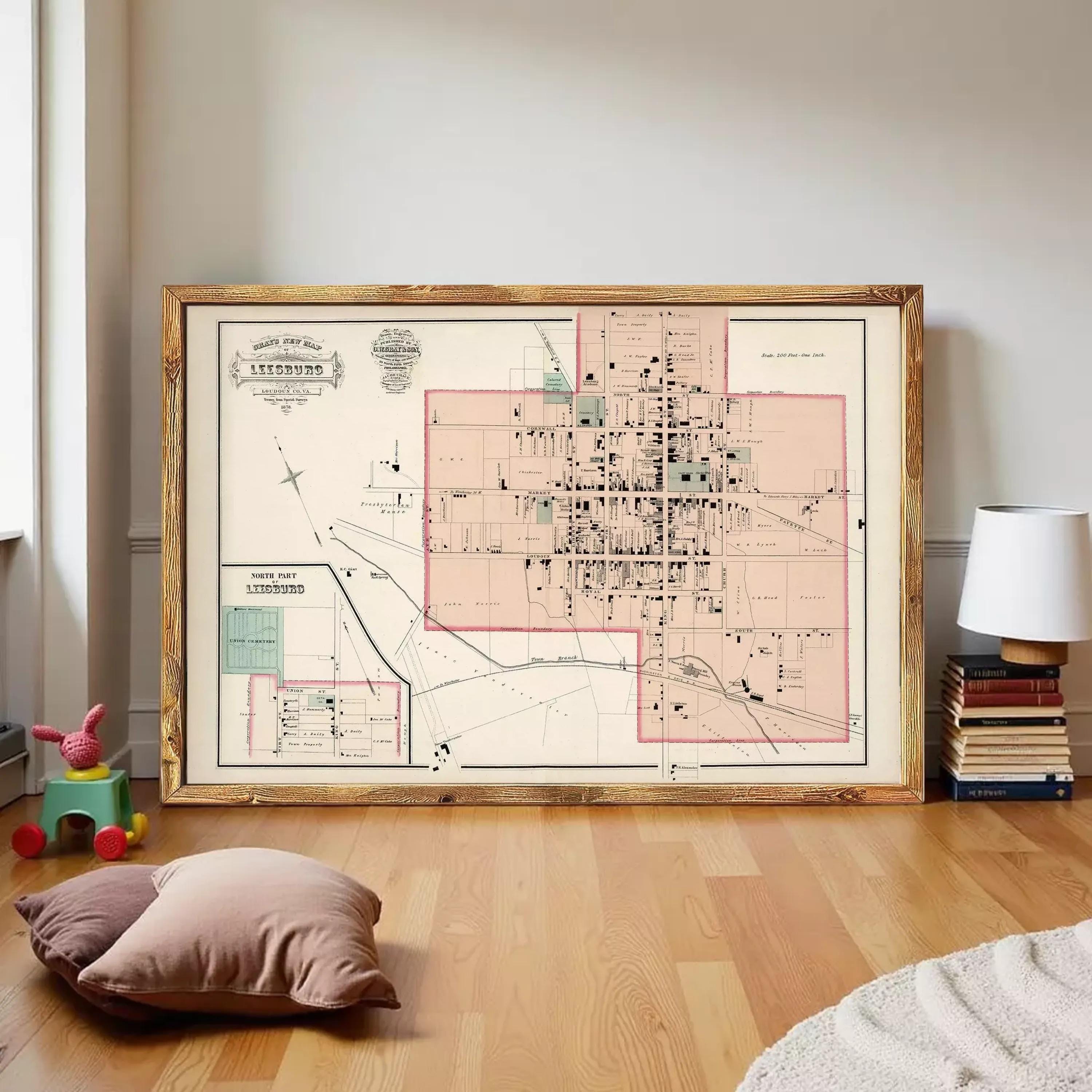 a large framed map of Leesburg, Virginia, with a wooden frame and a vintage design. The map is displayed on a wooden floor in a room with a lamp and some books.