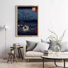 a cozy living room with a large framed artwork of a nighttime landscape hanging on the wall, featuring a dark blue background with a tree and glowing yellow lights. The room is furnished with a gray sofa, a wooden side table, and a black coffee table with a vase of greenery.