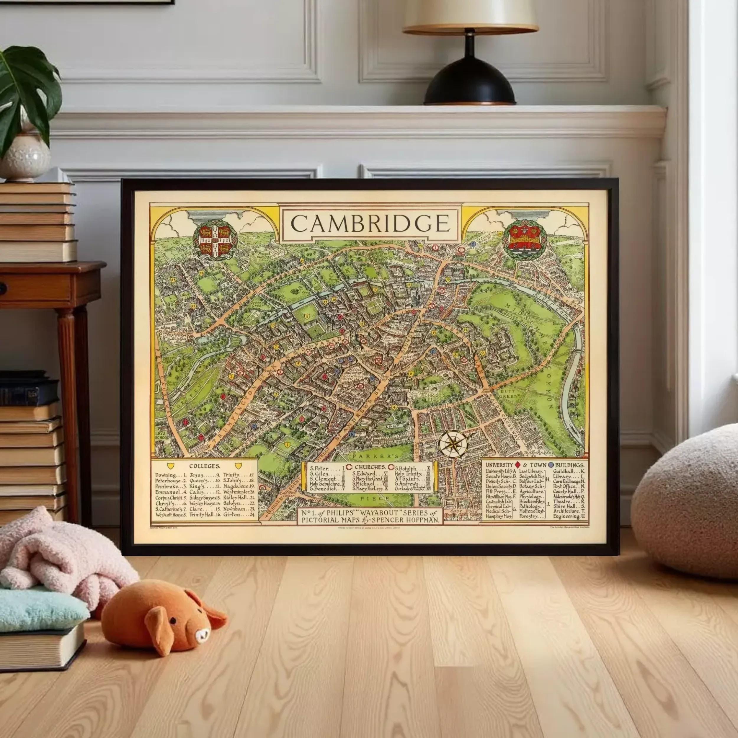 A framed map of Cambridge, England, displayed on a wooden floor in a room with a lamp and books.