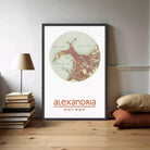 A framed map of the city of Alexandria, with the name "Alexandria" printed below it. The map is displayed on a wall, and there are some books and pillows visible in the foreground.