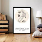 A framed poster of a map of New Zealand is displayed on a wall in a room with a bookshelf and a lamp.