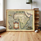 a large framed map of Africa, with various countries and regions clearly marked. The map is displayed on a wooden floor, with a stack of books and a lamp visible in the background.
