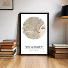 A framed map of Milwaukee, Wisconsin, with the city's name and coordinates displayed, is placed on a wooden floor next to a stack of books.
