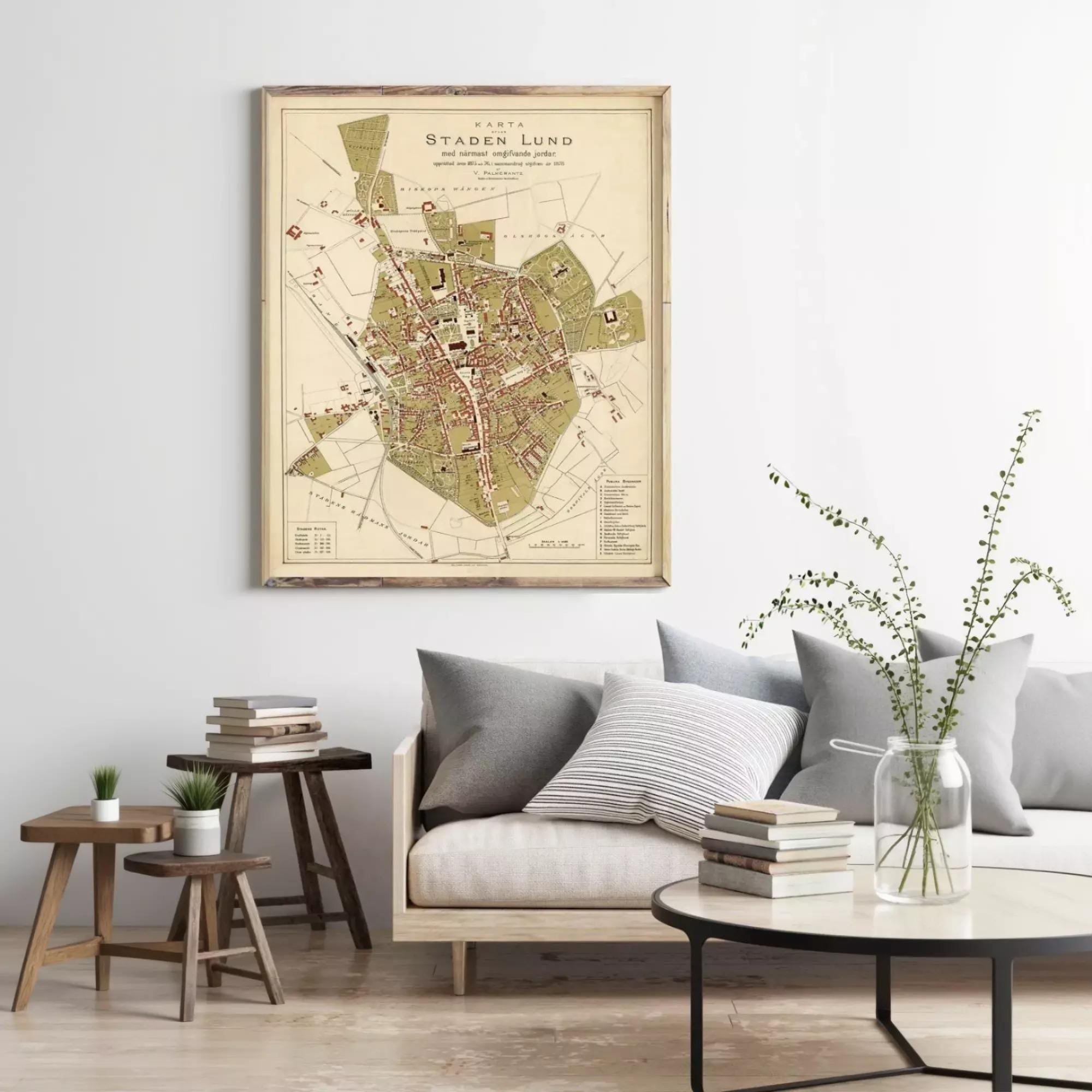 a living room with a large framed map of Staden Lund hanging on the wall, a wooden coffee table with books and a vase of greenery, and a couch with pillows.