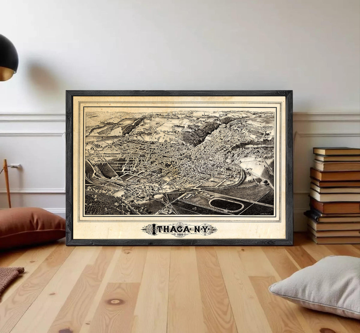 A framed black and white map of Ithaca, New York, displayed on a wooden floor in a room with books and pillows.