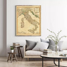 a living room with a large framed map of Italy hanging on the wall above a couch with pillows. There are also some books and a vase with greenery on the couch.