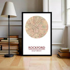 A framed map of Rockford, Illinois, with the text "ROCKFORD 42.16 16.89 W" printed on it, is displayed on a wooden floor next to a stack of books and a lamp.