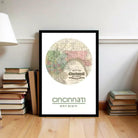 A framed map of Cincinnati, Ohio, with the text "Map of Cincinnati" and "Cincinnati, Ohio" printed on it, is displayed on a wooden floor next to a stack of books.