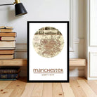 A framed map of Manchester, UK, with the text "Manchester" and "53°30'N 2°15'W" printed on it. The map is displayed on a wooden floor, with a stack of books and a lamp visible in the background.