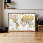 A large world map in a wooden frame is displayed on a wooden floor, surrounded by books and pillows.