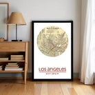 A framed vintage map of Los Angeles, with the text "Los Angeles" and "3,828 N, 1815 W" printed on it, is displayed on a wall next to a wooden shelf and a lamp.