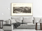 a large black and white framed picture of a countryside landscape hanging on a wall above a gray sofa with a plaid throw blanket.