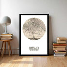 A framed map of Berlin, with a detailed illustration of the city's streets and landmarks, is displayed on a wall in a room with a stack of books and a lamp.