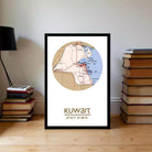 A framed map of Kuwait, with the country's borders and surrounding areas clearly visible. The map is placed on a wooden floor, with a stack of books to the left and a lamp to the right.