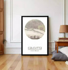 a framed poster of Calcutta, India, with a map of the city displayed on it. The poster is placed on a wooden floor in a room with a lamp and some books visible in the background.