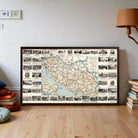The image is a large framed map of a region, likely a country or region, with various landmarks and locations marked on it. The map is displayed on a wooden floor in a room with a lamp and some books visible in the background.