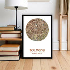 A framed map of Bologna, Italy, with the text "Bologna 44° 30' 11' 21"", placed on a wooden floor next to a stack of books and a lamp.