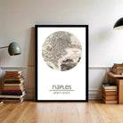 A framed map of Naples, Italy, with the text " Naples" and "40 50 50 14 15 e" printed on it, displayed on a wall in a room with books and a lamp.
