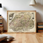 a large framed map of a mountainous region, with a wooden frame and a vintage appearance. The map is displayed on a wooden floor, surrounded by a stack of books and a lamp.