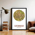 A framed map of Cambridge, UK, with the text "Cambridge" and "S12n 07O" printed on it, is displayed on a wall next to a stack of books and a lamp.