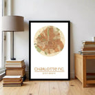A framed map of Charlotte, North Carolina, with the city's location and coordinates printed on it, placed on a wooden floor next to a stack of books and a lamp.