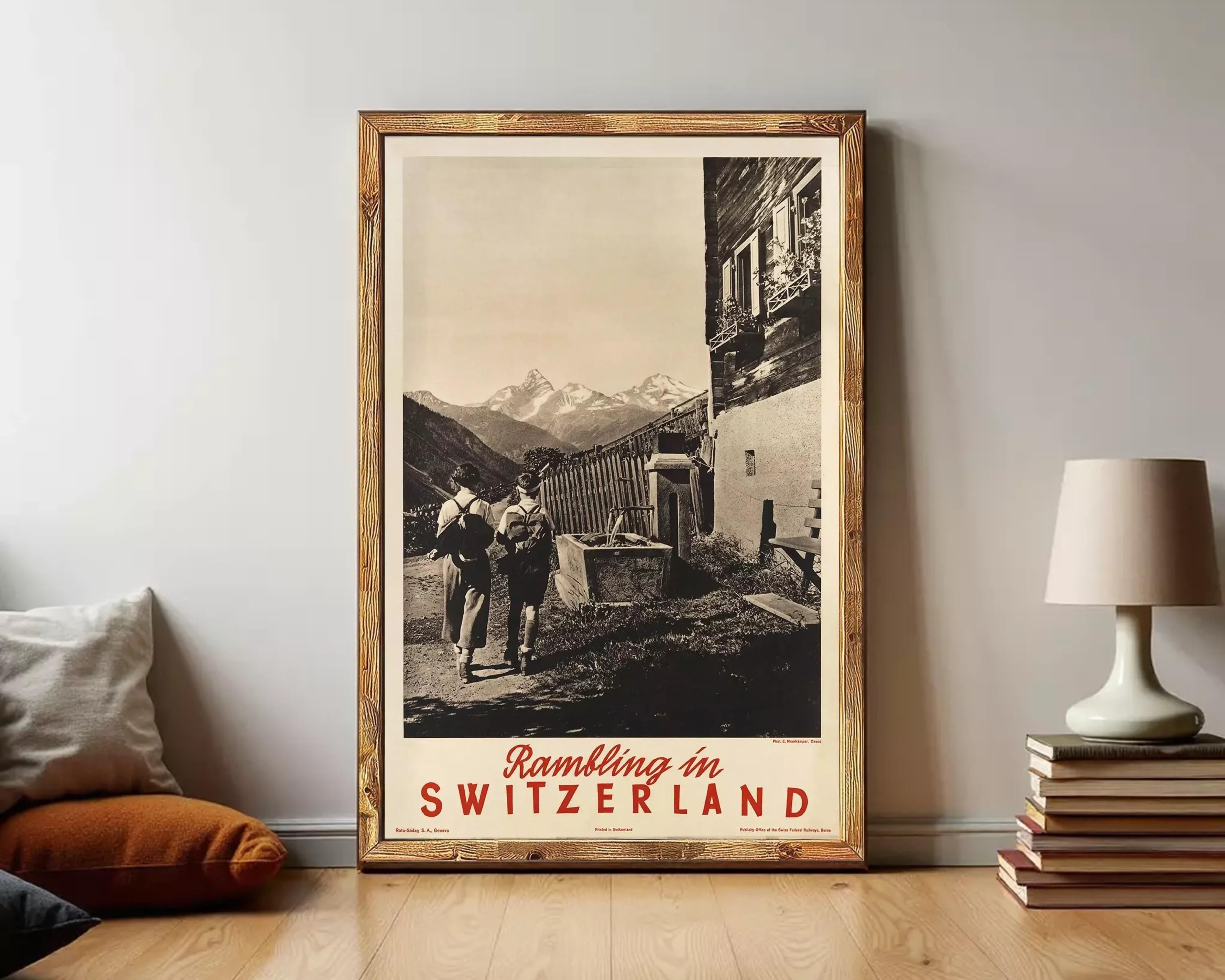 The image is a framed vintage travel poster depicting a couple walking on a path in a mountainous landscape, with a Swiss chalet in the background.