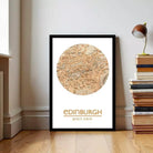 A framed map of Edinburgh, Scotland, with the city's name and location printed on it, is displayed on a wall in a room with a stack of books and a lamp.