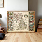 A large framed map of the United Kingdom, featuring intricate illustrations and details, is displayed on a wooden floor in a room with books and other decor.