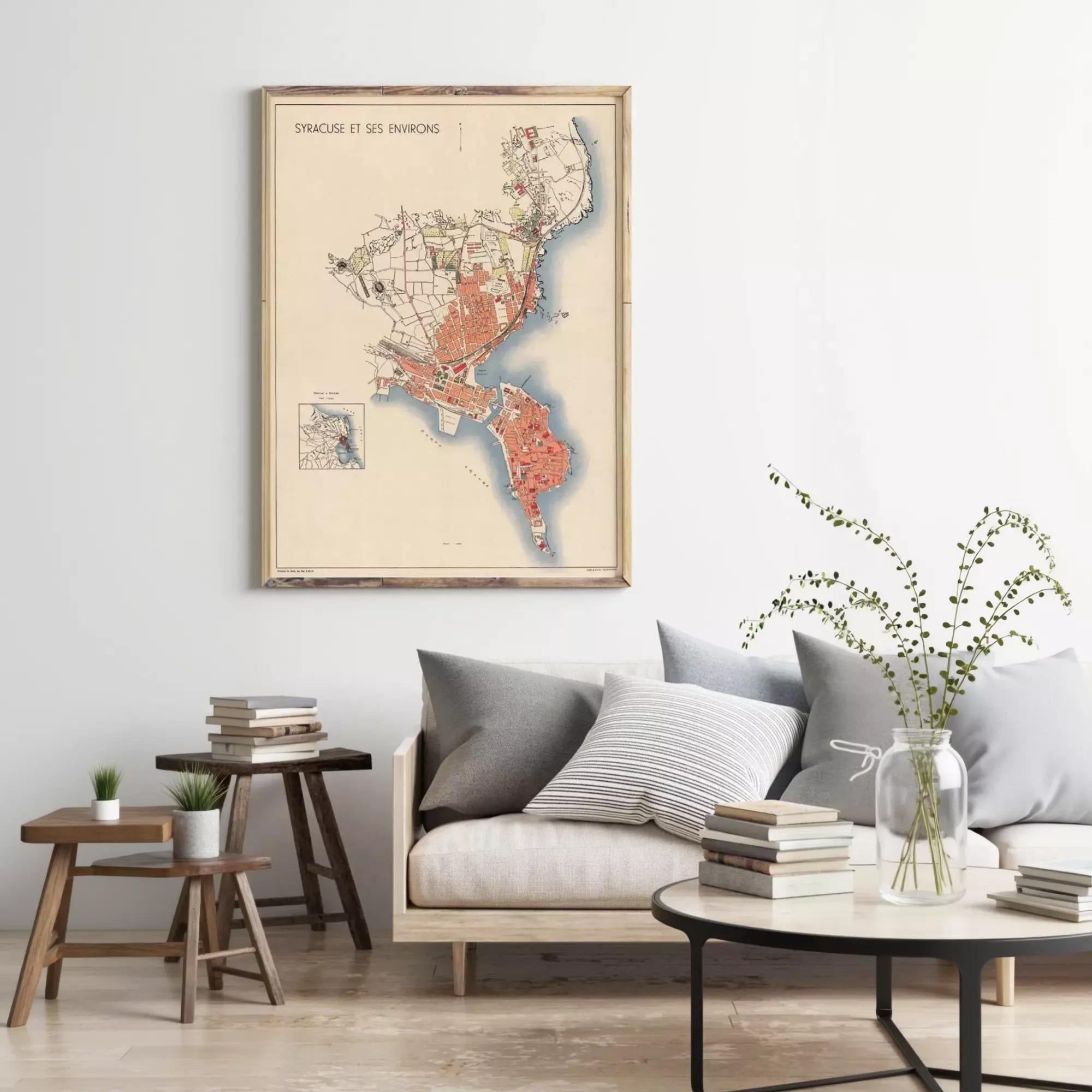 a living room with a framed map of a city hanging on the wall, a couch with pillows, a coffee table with books and a vase of greenery, and a small side table with a plant.