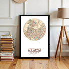 A framed map of Ottawa, Canada, with the city's name and location printed on it, is displayed on a wooden floor next to a stack of books and a lamp.