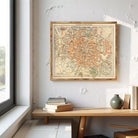 a framed map of a city hanging on a wall, with a wooden table below it and some books and a vase on the table.