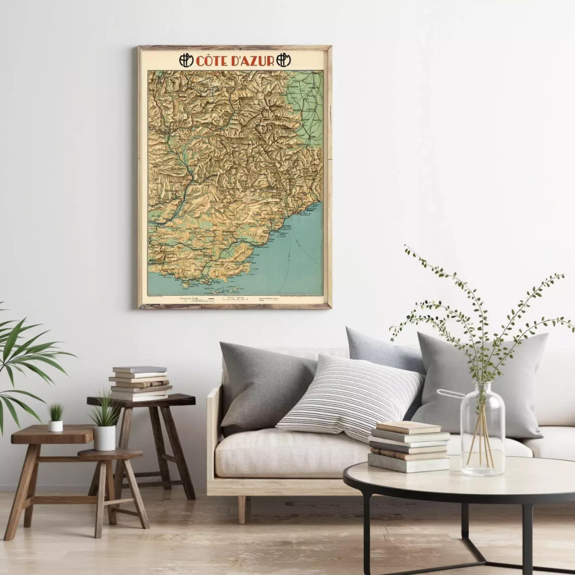 a living room with a large framed map of Cote d'Azur hanging on the wall above a couch with pillows. The room also includes a coffee table with books and a vase of greenery.