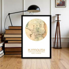 A framed map of Plymouth, Massachusetts, with the city's location and surrounding area, is displayed on a wooden floor in a room with a lamp and bookshelves.