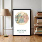 A framed map of Utica, New York, with a lamp and a bookshelf in the background.