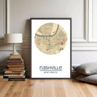 A framed map of Nashville, Tennessee, with the text " Nashville 36°10'11" 86°47'7"", placed on a wooden floor next to a stack of books and a lamp.